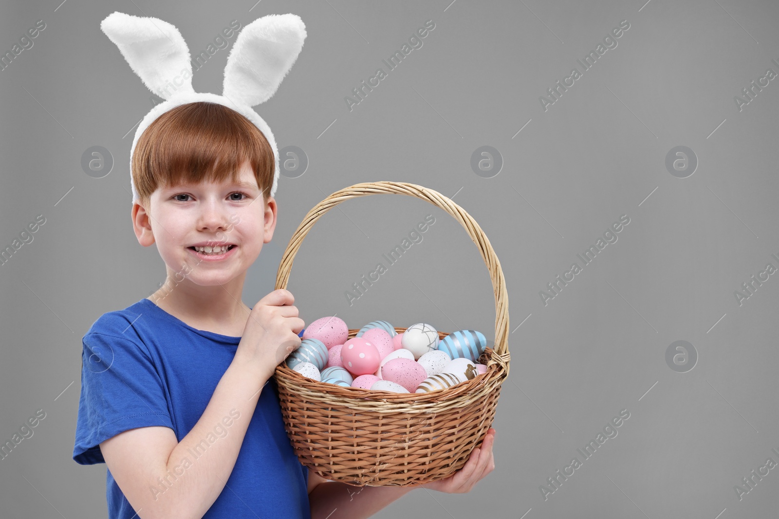 Photo of Easter celebration. Cute little boy with bunny ears and wicker basket full of painted eggs on grey background. Space for text
