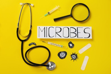 Photo of Card with word Microbes, magnifying glass, syringe, stethoscope and thermometer on yellow background, flat lay