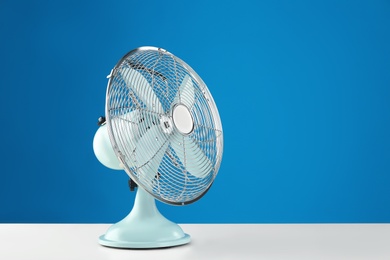Photo of Electric fan on white table against blue background, space for text. Summer heat