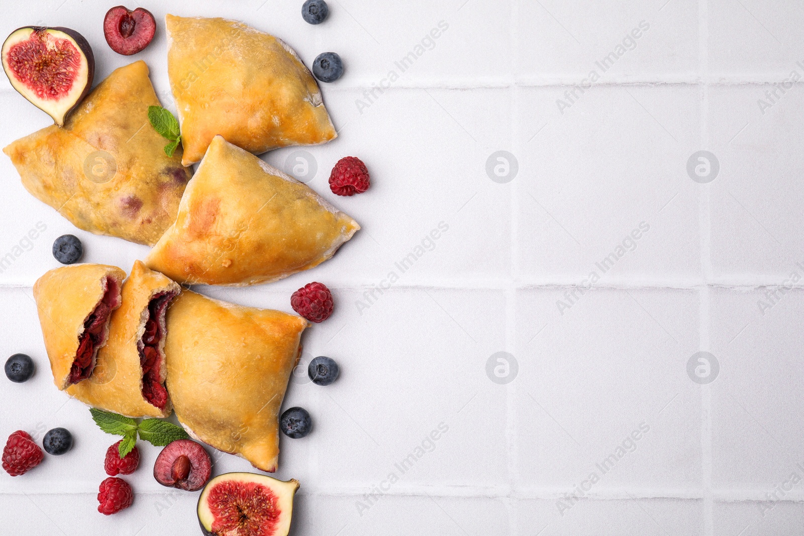 Photo of Delicious samosas, berries, mint leaves and fig on white tiled table, flat lay. Space for text