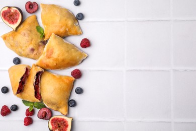 Photo of Delicious samosas, berries, mint leaves and fig on white tiled table, flat lay. Space for text