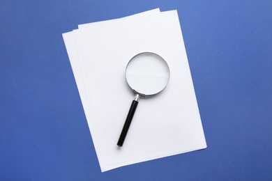 Magnifying glass and paper sheets on blue background, top view