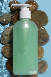 Bottle of face cleansing product and stones in water against light blue background, flat lay