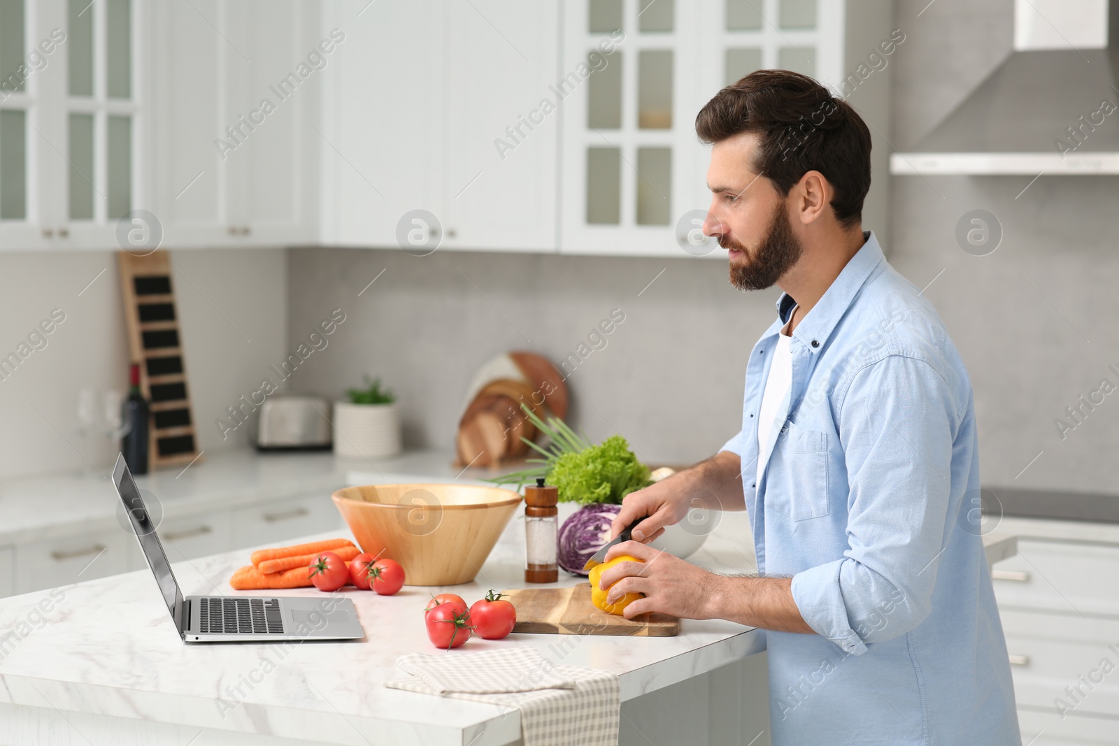 Photo of Man making dinner while watching online cooking course via laptop in kitchen