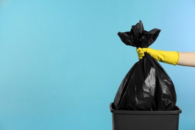 Photo of Janitor in rubber glove holding trash bag full of garbage over bucket on light blue background, closeup. Space for text