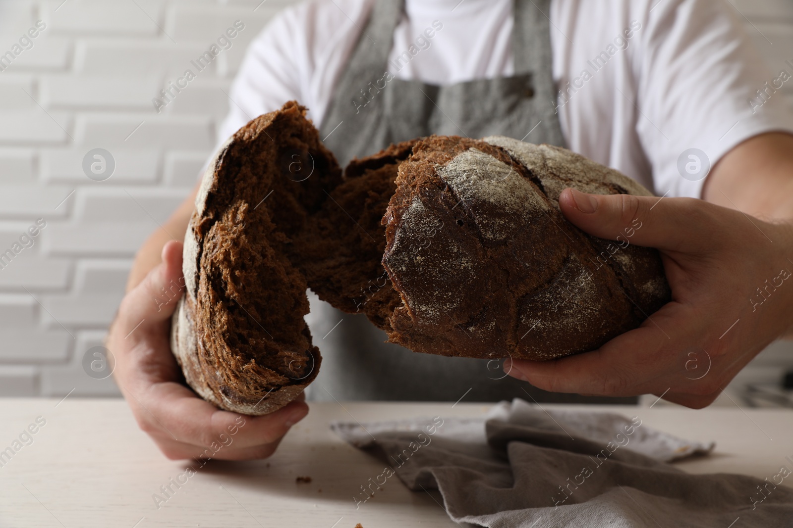 Photo of Man breaking loaf of fresh bread at white table near brick wall, closeup