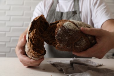 Photo of Man breaking loaf of fresh bread at white table near brick wall, closeup