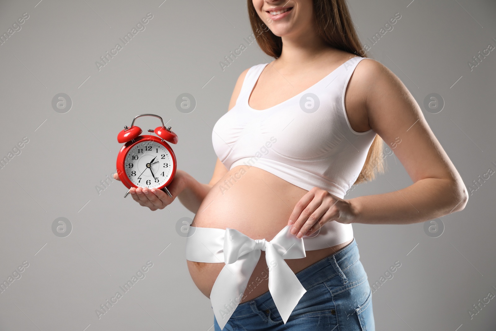 Photo of Young pregnant woman with alarm clock and bow on grey background, closeup. Time to give birth