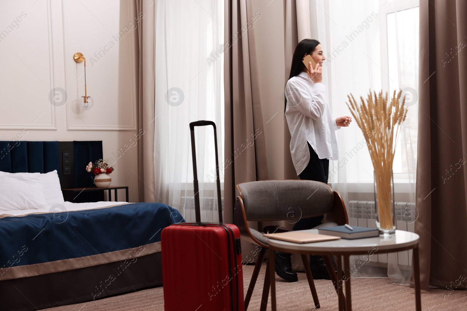 Photo of Beautiful businesswoman talking on phone near window in hotel room