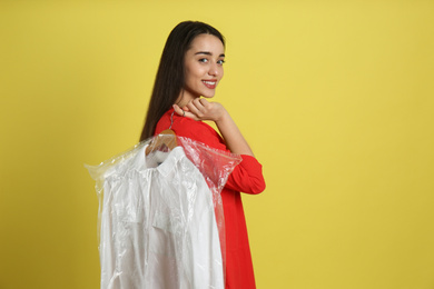 Photo of Young woman holding hanger with shirt in plastic bag on yellow background. Dry-cleaning service