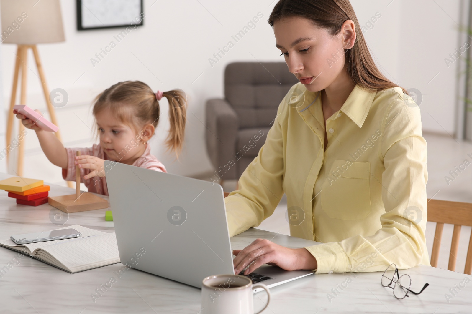 Photo of Woman working remotely at home. Mother using laptop while daughter playing at desk