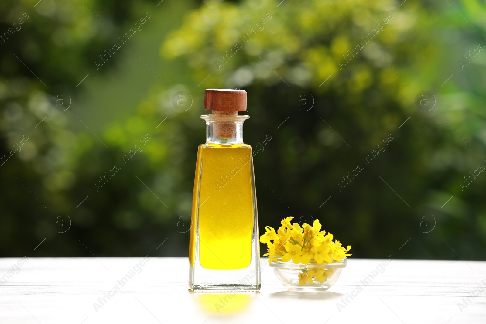 Photo of Rapeseed oil in glass bottle and yellow flowers on white wooden table outdoors