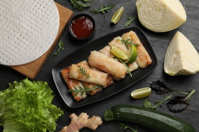 Photo of Tasty fried spring rolls, arugula, sauce and other products on dark textured table, flat lay