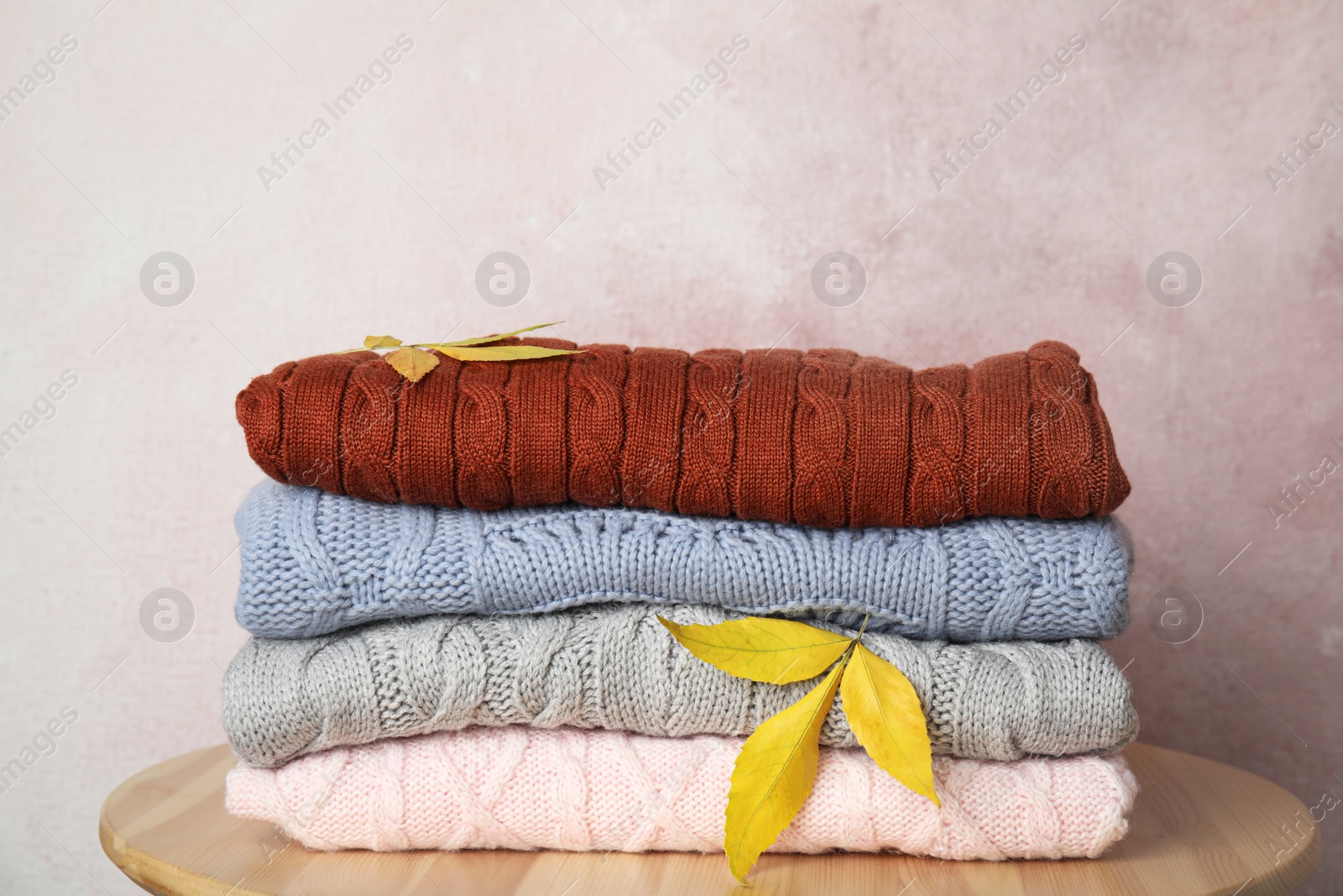 Photo of Stack of warm knitted clothes and autumn leaves on wooden table against light pink background