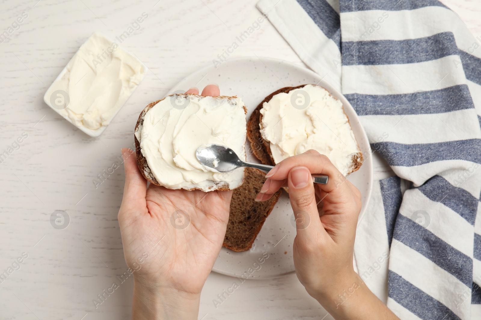 Photo of Woman spreading cream cheese onto bread at white wooden table, closeup