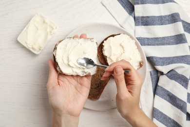 Woman spreading cream cheese onto bread at white wooden table, closeup