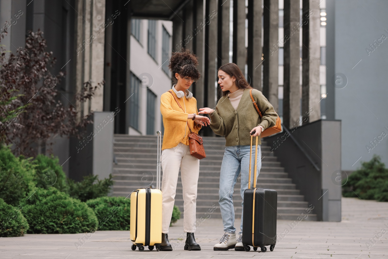 Photo of Being late. Worried women with suitcases looking at watch near building outdoors