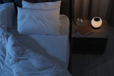 Photo of Nightlight, alarm clock and book on bedside table near bed indoors, above view