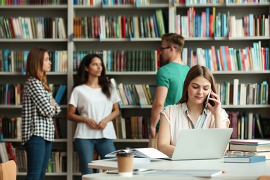 Photo of Young woman talking on phone at table and people standing near bookshelves in library