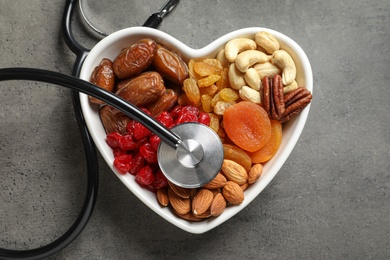Photo of Heart shaped bowl with dried fruits, nuts and stethoscope on grey background, top view