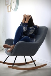 Depressed young woman in rocking chair near light wall