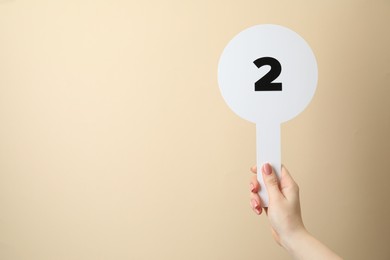 Woman holding auction paddle with number 2 on beige background, closeup. Space for text