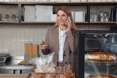 Photo of Happy business owner talking on phone at cashier desk in bakery shop
