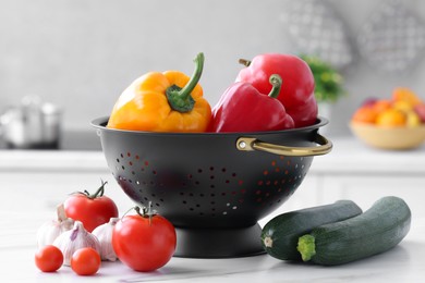 Black colander and different vegetables on white marble table in kitchen