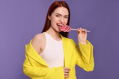 Photo of Stylish woman with red dyed hair biting lollipop on purple background