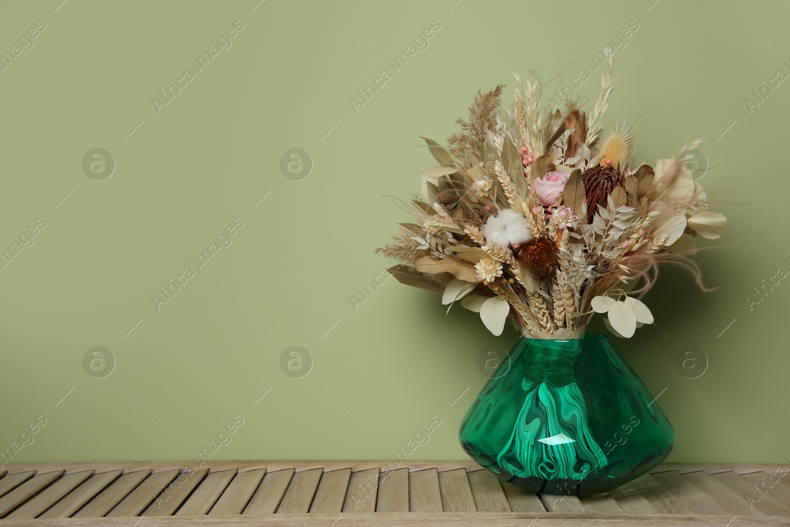 Photo of Beautiful dried flower bouquet in glass vase on wooden table near olive wall. Space for text