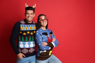 Photo of Couple in Christmas sweaters, deer headband and party glasses on red background, space for text
