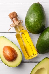 Glass bottle of cooking oil and fresh avocados on white wooden table, flat lay