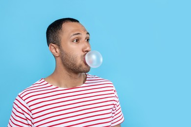 Young man blowing bubble gum on light blue background. Space for text