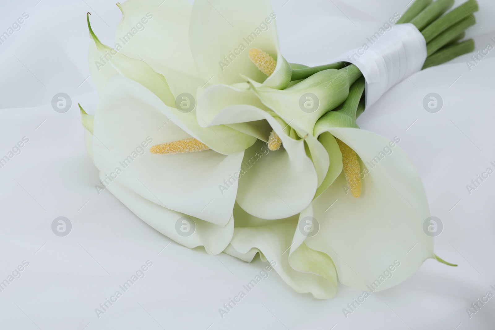 Photo of Beautiful calla lily flowers tied with ribbon on white fabric, closeup