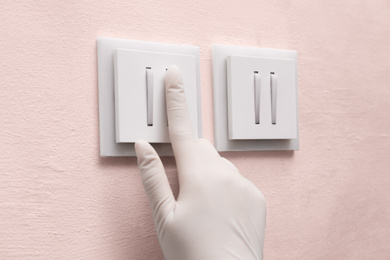 Photo of Woman in protective gloves pressing button of light switch indoors, closeup