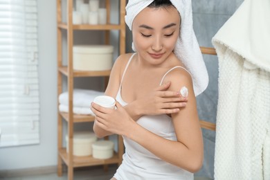 Photo of Beautiful young Asian woman applying body cream on shoulder in bathroom, space for text