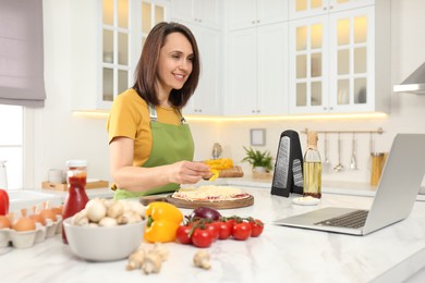 Photo of Woman making pizza while watching online cooking course via laptop in kitchen