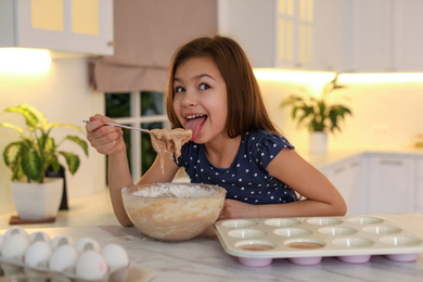 Photo of Cute little girl licking raw dough from spoon in kitchen. Cooking food