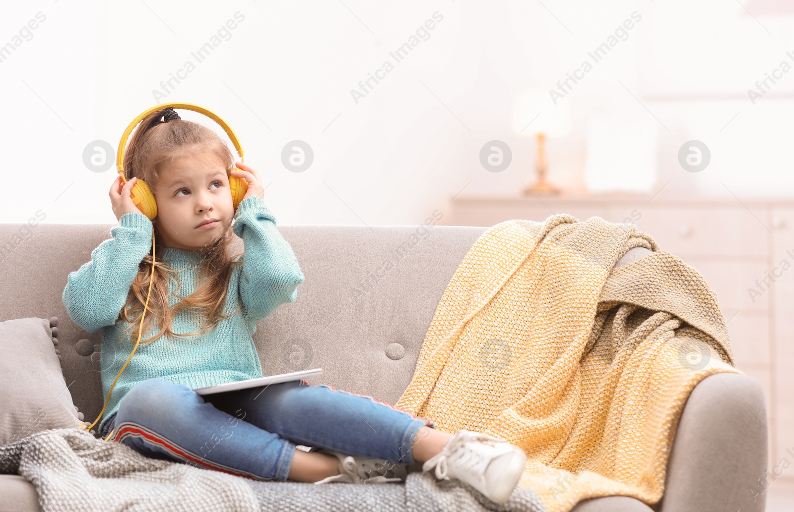 Photo of Cute little girl with headphones and tablet on sofa at home. Space for text