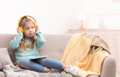 Cute little girl with headphones and tablet on sofa at home. Space for text