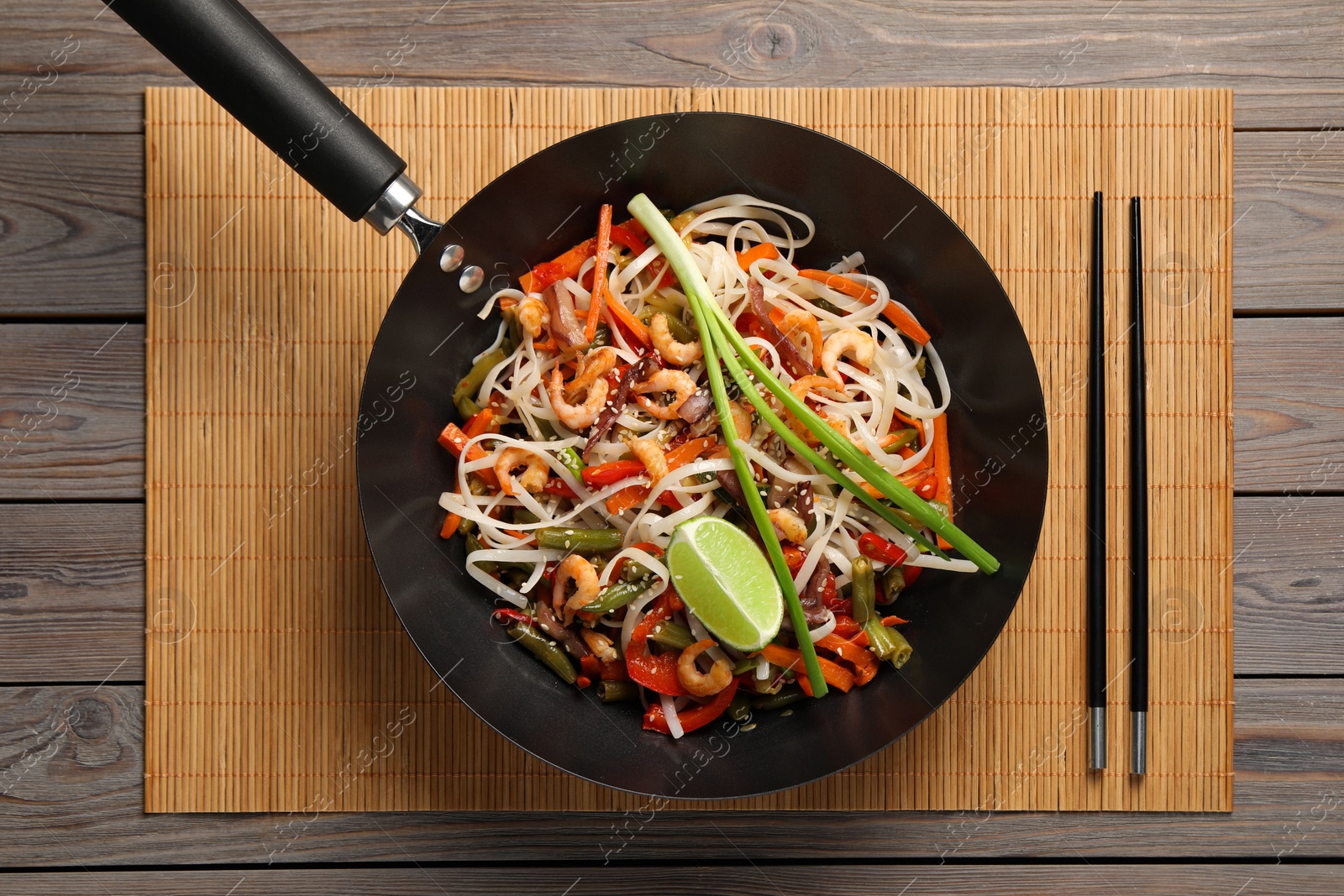 Photo of Shrimp stir fry with noodles and vegetables in wok on wooden table, top view