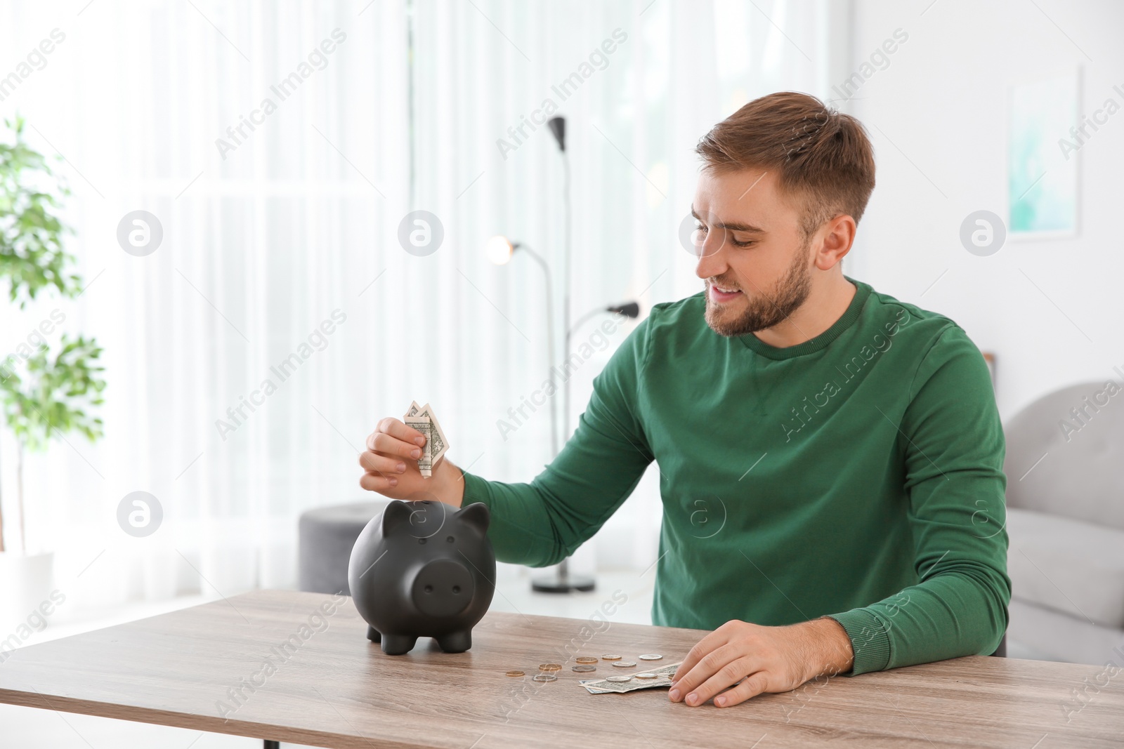 Photo of Young man putting coin into piggy bank at table indoors. Space for text