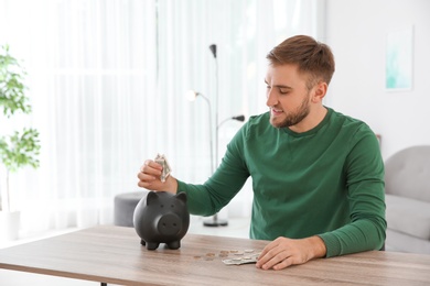 Photo of Young man putting coin into piggy bank at table indoors. Space for text