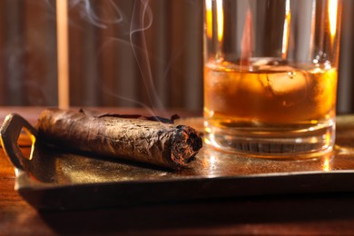 Tray with smoldering cigar and whiskey on wooden table, closeup
