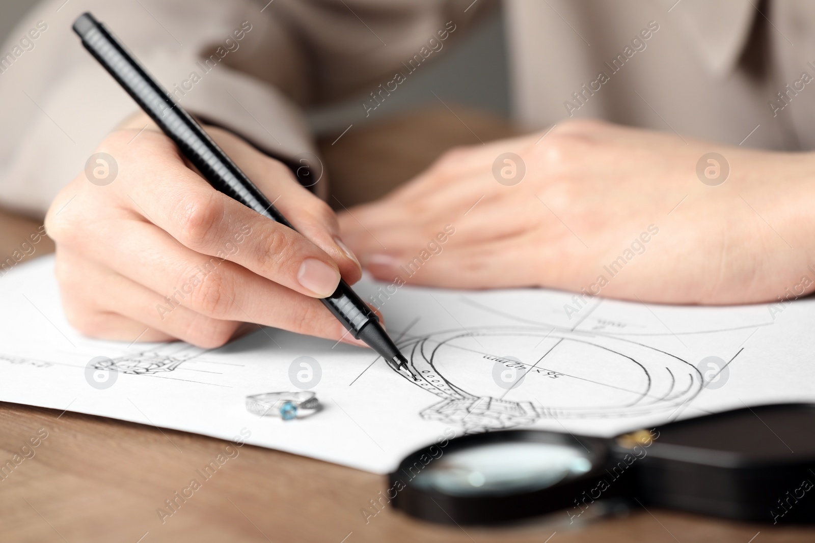 Photo of Jeweler drawing sketch of elegant ring on paper at wooden table, closeup