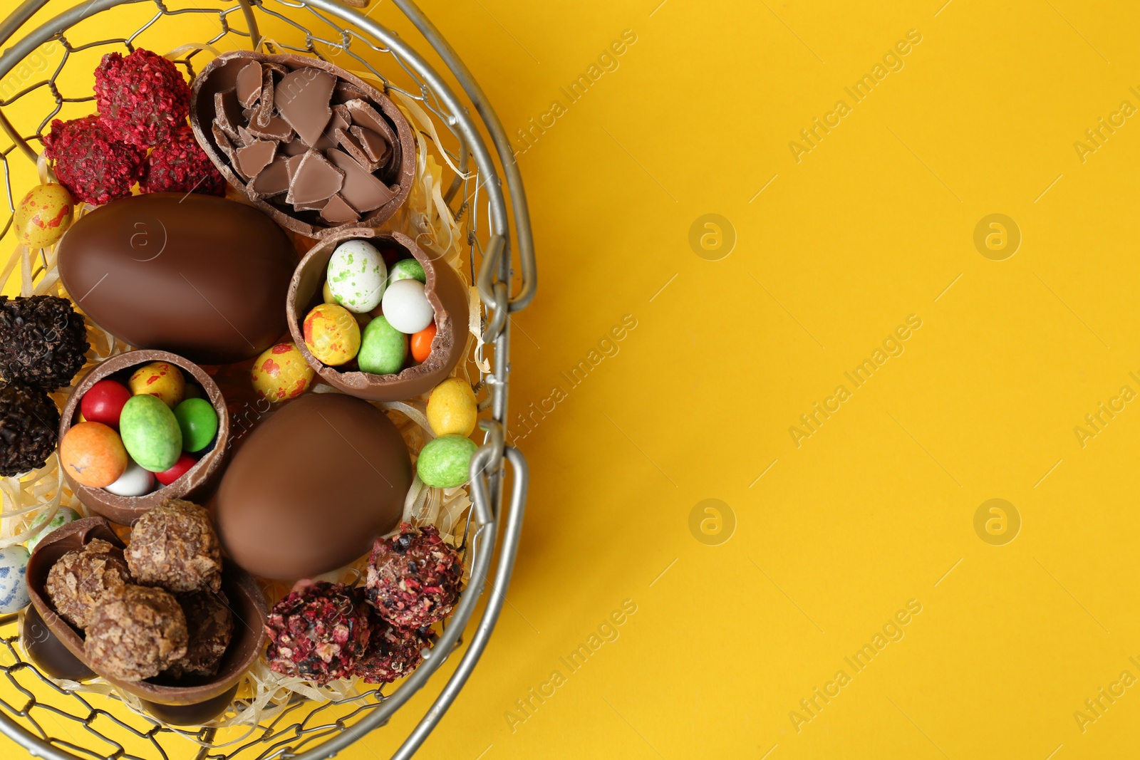 Photo of Basket with chocolate eggs and candies on yellow background, top view. Space for text