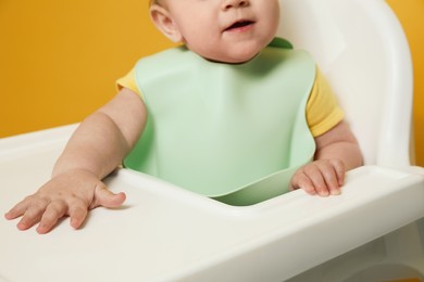 Photo of Cute little baby wearing bib in highchair on yellow background, closeup