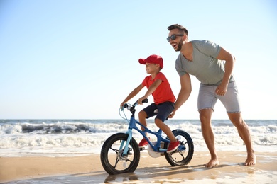Photo of Happy father teaching son to ride bicycle on sandy beach near sea