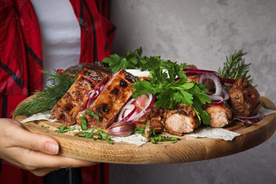 Photo of Woman holding wooden board with delicious roasted meat and herbal on grey background, closeup