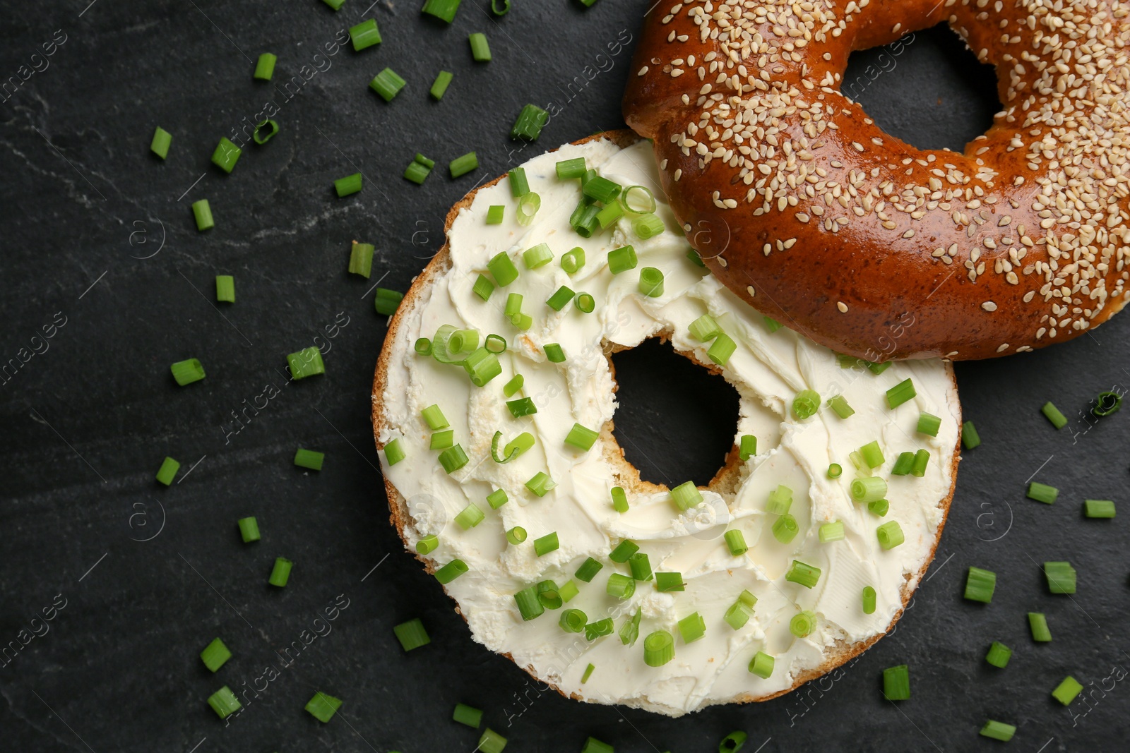 Photo of Delicious bagel with cream cheese and green onion on black table, flat lay